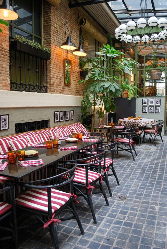 Cozy interior of Fellina restaurant in Madrid featuring warm vintage decor, exposed brick walls, and lush green plants hanging from the ceiling.