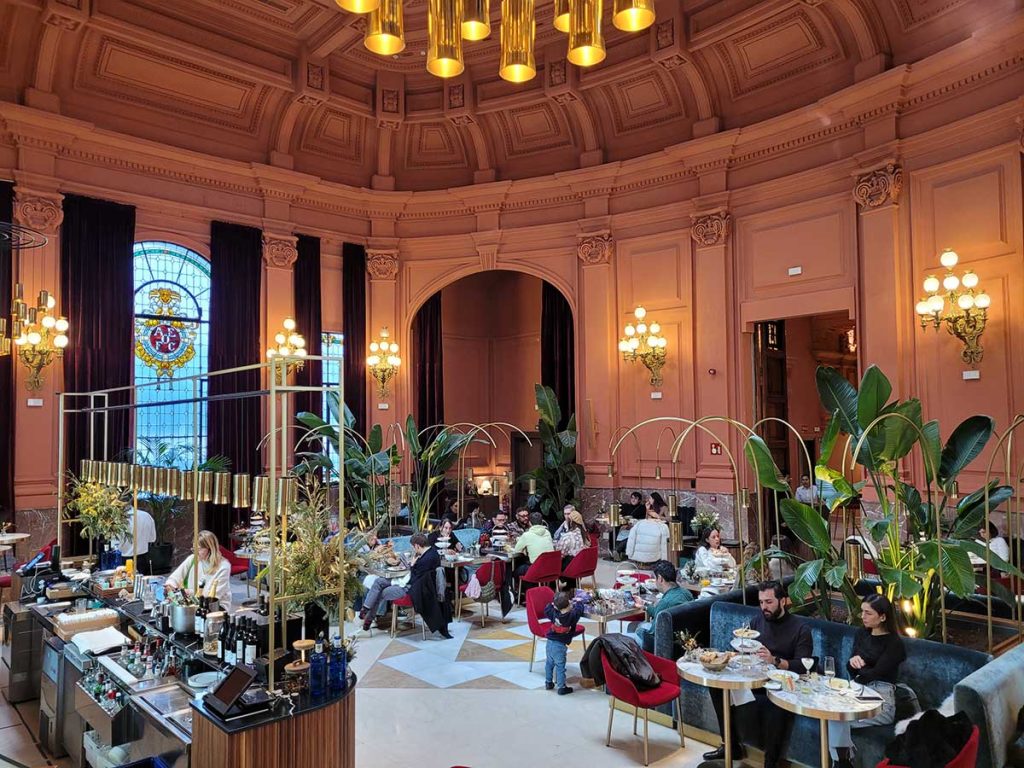 Interior of Maché Restaurant in Madrid featuring high ceilings with classic moldings, large stained-glass windows, velvet seating, and warm spherical pendant lights reflecting off polished floors.