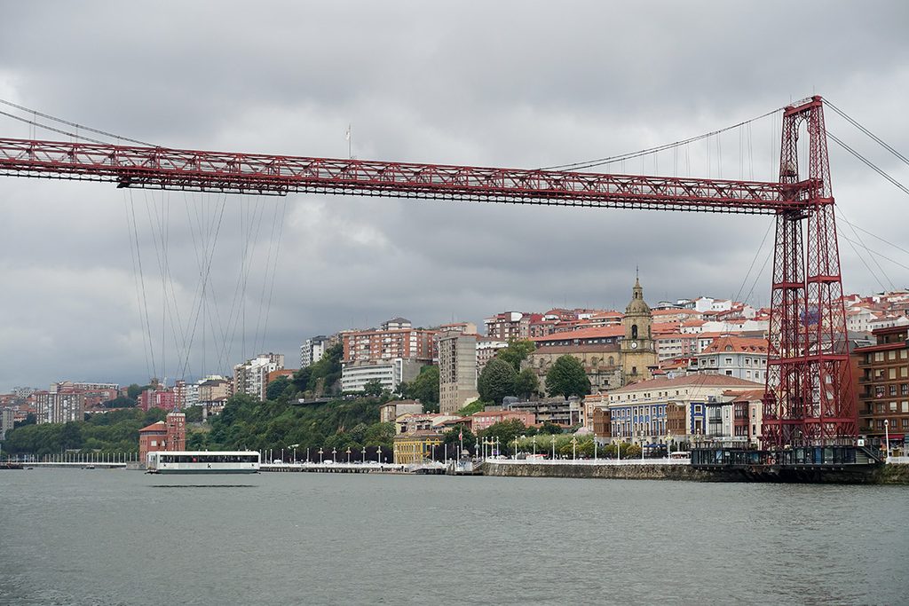 A wide shot of the historic, industrial iron Vizcaya Bridge stretching across the river, with its suspended transport gondola carrying passengers over the water